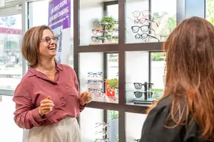 A woman wearing glasses is talking to another woman in a room with a glass wall and display of eyeglasses