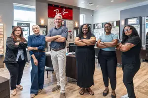 Six people are posing for a photo in an optical store with a Ray-Ban sign in the background