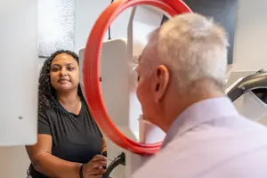 An adult man is getting his eyes checked by a woman in a black shirt in a room with a red circular mirror.