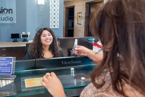 A woman is smiling while sitting at a reception desk and another woman is handing her something in a small bottle