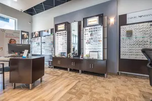 A room with wooden flooring and a monitor on a desk, there are cabinets, glass cabinets with different kinds of eyeglasses, and a surveillance camera on the ceiling.