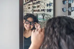 A woman with glasses is adjusting her glasses in front of a mirror in a store