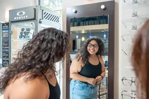 A woman is looking at her reflection in a mirror, smiling, with another woman looking at her from behind, and a display case with Oakley sunglasses in the background.