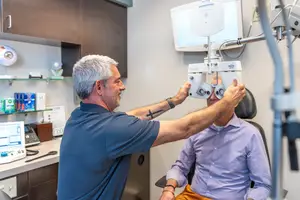 A man in a blue shirt adjusts a machine over the eye of a man in a purple shirt sitting in a chair.