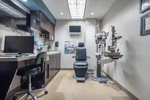 An empty optometrist examination room with a black desk and chair, a computer monitor, a sink, and an eye examination machine in the corner.