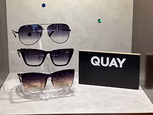Three pairs of Quay sunglasses are displayed on a clear holder next to a black box with the word Quay on it.