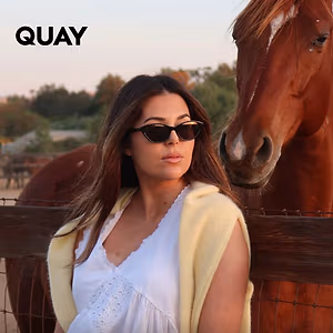 A woman in a white top and sunglasses stands near a horse in a fenced area