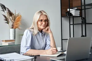 A smiling woman wearing glasses sits at a desk with a laptop, notebook, and pen, surrounded by books and a flower vase.
