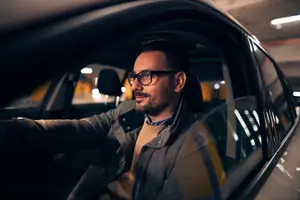 A man with glasses sits in a car parked in a parking garage, looking sideways with a smile.
