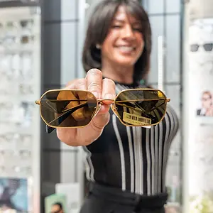 A woman wearing a striped black and white dress is smiling and holding a pair of Prada sunglasses with golden frames and lenses, and the reflection of her hand and the glasses is visible on the lens.