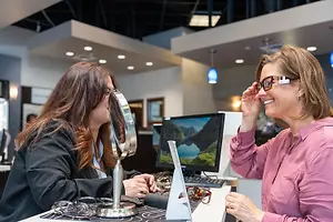 Two women, one wearing glasses and smiling, while the other is looking at something on a desk in a store.