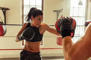 A woman wearing a black sports bra and boxing gloves is punching a red punching bag with her right hand in a boxing gym with a man wearing red and black boxing gloves standing on the side.