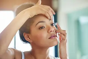 A woman is putting in her contact lens while standing in front of a mirror