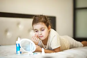 A young girl is lying on the bed while applying a facial cream on her face with her right hand