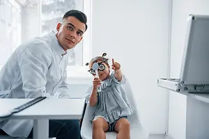 A man wearing a white lab coat is examining a young girl's eyes with a magnifying glass in an office room.