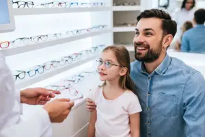 A man and a girl are in an optical store, looking at glasses, with a person holding a pair of glasses, a cabinet filled with glasses, and a monitor on the left side.