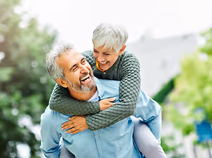 An elderly man and woman are outside on a sunny day, the woman is sitting on the man's shoulders and smiling, the man is looking at her and smiling back