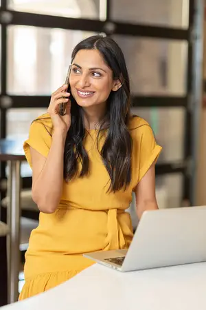 A smiling woman is on a phone call while sitting in front of a laptop