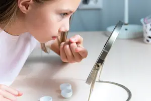 A young girl is sitting in front of a desk, holding a contact lens and looking at herself in a mirror