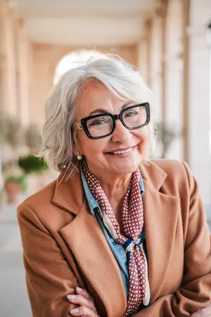 A smiling woman wearing glasses and a scarf is standing in front of a building with a pillar