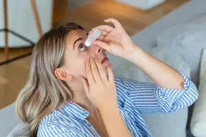 A woman is applying eye drops to her eye while sitting on a couch