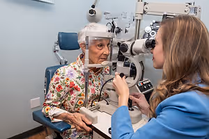 An elderly woman with white hair is getting her eye checked by a young woman in a blue coat in a clinic room with a white wall and a blue chair.