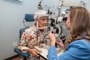An elderly woman with white hair is getting her eye checked by a young woman in a blue coat in a clinic room with a white wall and a blue chair.
