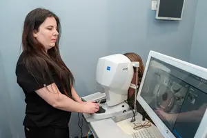 A woman is adjusting the settings on an eye exam machine while another woman is sitting in front of the monitor in a blue room.