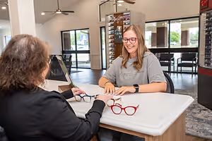 A woman is sitting at a desk in an office store and smiling while another woman is holding a pair of glasses and they are both looking at the glasses on the desk.