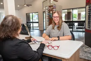 A woman is sitting at a desk in an office store and smiling while another woman is holding a pair of glasses and they are both looking at the glasses on the desk.