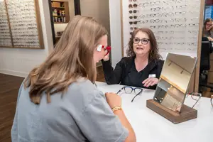 A woman is looking at herself in a mirror while another woman is sitting next to her. They are both looking at each other and smiling. Behind them are shelves with different kinds of eyeglasses. There is a wooden mirror on the table with a pair of eyeglasses on it.