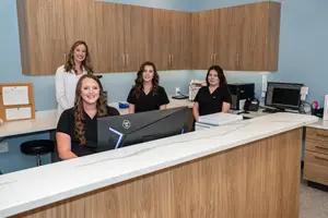 Four women smiling and posing for a photo in an office with wooden cabinets, a monitor, and a counter