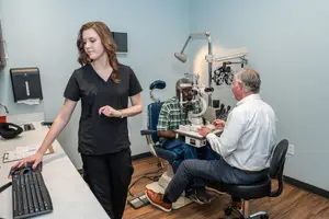 A woman stands next to a table with a keyboard while an elderly man sits in a chair next to an eye doctor who is examining a patient