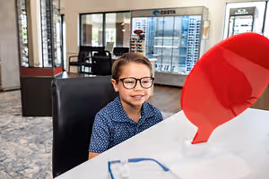 A boy wearing glasses is sitting on a chair in front of a desk with a red object in the optical shop.