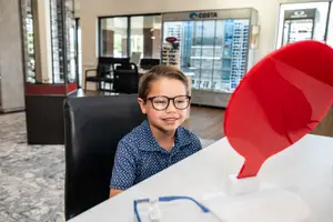 A boy wearing glasses is sitting on a chair in front of a desk with a red object in the optical shop.