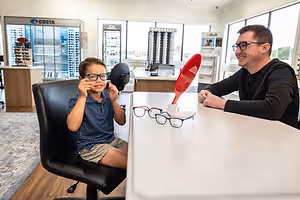 A young boy wearing glasses is sitting on a chair and looking at a man wearing glasses in an optical shop