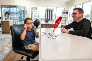 A young boy wearing glasses is sitting on a chair and looking at a man wearing glasses in an optical shop
