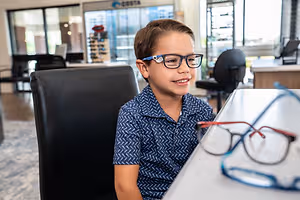 A young boy with glasses is smiling while sitting at a desk in an office with a blurred background.