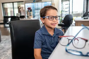 A young boy with glasses is smiling while sitting at a desk in an office with a blurred background.