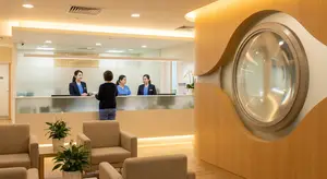 Three women wearing blue uniforms work at a glass fronted reception desk while a woman stands nearby, and a large curved mirror hangs from the wall
