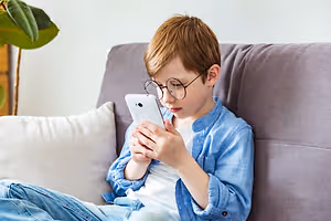 A boy sitting on a couch with a white phone in his hands, looking at the screen.