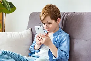 A boy sitting on a couch with a white phone in his hands, looking at the screen.