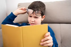 A boy sitting on a couch wearing glasses is holding a book with both hands and appears to be reading it while his other hand is on his glasses.