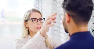 A woman wearing glasses is showing a man how to wear them correctly while standing in front of a glass window.