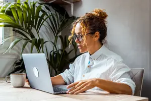 A woman with curly hair and glasses is working on a laptop on a table with a coffee mug, a plant, and a white wall in the background.
