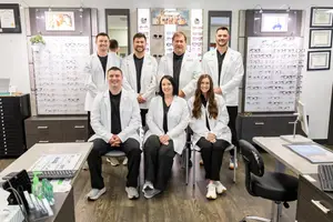 A group of people in white coats and sneakers are posing for a photo in an optical shop with cabinets, shelves, and monitors in the background.