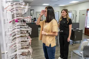 Two women are standing in front of a row of glasses in an optician's shop, with one woman raising her hand as if trying on glasses and the other woman looking at her, and behind them are a monitor, a desk with a computer, and a mirror on the wall.