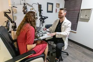A man in a white coat is examining a woman's eyes with an ophthalmoscope in an office.