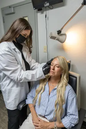 A woman is sitting in a chair while another woman is standing beside her, holding her eyes, and appears to be doing some treatment with her eyes closed