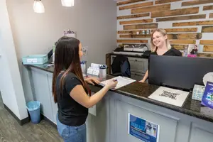 An adult woman is standing in front of the reception desk and talking to another woman who is standing behind the desk. The woman in front is holding a pen and a notepad. On the desk are a trash bin, a storage box, a cup, a pen holder, a monitor, and a poster.